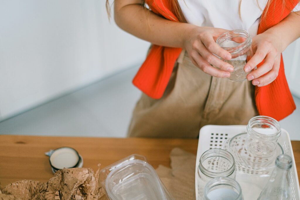 Crop anonymous female in casual clothes standing with glass jar near table while sorting out paper plastic and glass rubbish for recycling